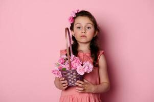 noble pequeño niña con Rizado pelo y hermosa azul ojos, participación cesta de rosado rosas, mirando perspicazmente a cámara foto