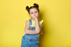 adorable pequeño niño niña en pie con brazos doblada, mirando pensativo, aislado terminado amarillo color antecedentes. inteligente niños foto