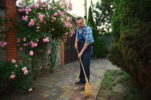 lleno longitud retrato de un jardinero hombre utilizando escoba, barre hojas y flor pétalos en el patio de un privado casa foto