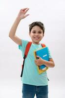 Emotional happy smart schoolboy greeting waving hand, smiling at camera, carrying orange backpack and school supplies photo