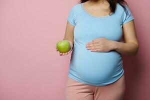 Midsection of pregnant expectant woman putting hand on her belly, holding a green apple, isolated over pink background. photo