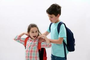 Caring teen boy puts on a backpack on his younger sister back, preparing her to the school, isolated on white background photo