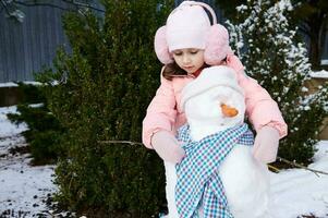 encantador pequeño niño niña pone en un azul chal en un muñeco de nieve, durante activo invierno ocio juegos en el Nevado patio interior foto