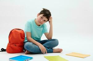 Puzzled doubtful pensive teenager school boy, scratches his head, sitting next to arranged textbooks and orange backpack photo