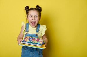 alegre niño niña con gracioso peinado, expresando asombro a cámara, que lleva libros y lápiz caso, amarillo fondo foto