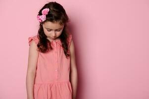 Sweet baby girl with pink rosed in hair, wearing stylish dress, looking down, posingover pink color background photo