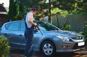 Full length portrait of a service man in blue work uniform, manually washing car with high pressure water jet outdoors. photo