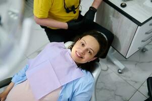 retrato de un joven embarazada mujer en dental silla, sonriente mirando a cámara durante un dentista cheque arriba. ver desde encima foto