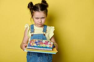 Emotional primary school student girl looking at camera with angry facial expression, holding pencil case and textbooks photo