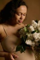 Close-up gorgeous gravid woman in beige lingerie, holding bouquet of blooming lilacs. Pregnancy fashion. Body positivity photo