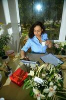 Beautiful woman florist, event manager taking sip of aroma coffee, checking orders for floral arrangement in flower shop photo