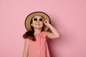 dañoso pequeño niño niña vistiendo Gafas de sol, Paja sombrero y verano vestido de verano, sonrisas mirando a cámara aislado en rosado foto