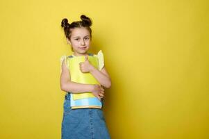 Cute kid girl, primary school student, thumbing up and smiling looking at camera, holding textbooks, isolated on yellow photo