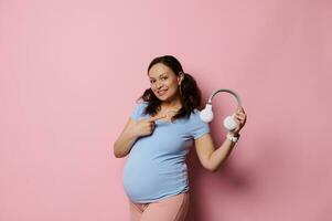 Cheerful pregnant woman in blue t-shirt, smiles and points finger aside wireless headphones, isolated on pink background photo