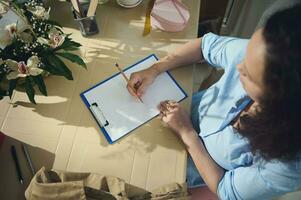 Directly above female entrepreneur florist holding a pencil, writing notes on clipboard in flower shop. Copy ad space photo