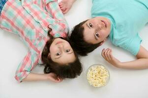 View from above of adorable kids in casual clothes lying on white background with a bowl of popcorn, looking at camera photo