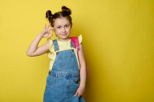 Happy primary school student girl, first grader showing OK sign with hand, smiling looking at camera, yellow background photo