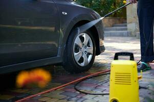 Close-up high pressure water cleaner as seen spraying on car wheel. Man washing car outdoors. Automobile service concept photo