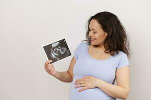 retrato de un contento embarazada mujer conmovedor barriga, participación ultrasonido imager Oh su bebé, aislado terminado blanco antecedentes foto