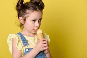 Close-up of little child girl licking scoop of ice cream in waffle cone, looking aside a copy space on yellow background photo