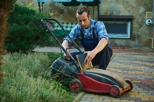 Hispanic gardener in blue work uniform, taking the cut grass out of the filter, recharging the lawn mower in courtyard photo