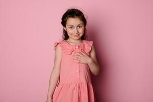 Beautiful shy little child girl in elegant pink dress, smiling cutely looking at camera, isolated on pink background photo
