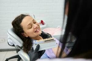 Dentist placing tooth color chart, over a smiling woman's teeth, choosing the shade of veneers, according to Vita scale. photo