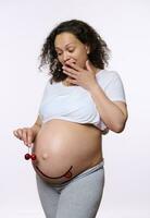 Amazed pregnant woman holds sweet cherries near hungry emoji showing tongue, created on her naked belly. Healthy eating photo