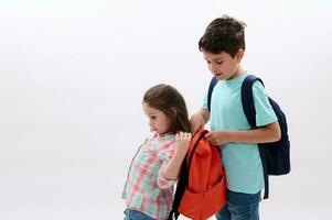 Caring teen boy older brother helps his younger sister to put on a backpack on back, isolated on white studio background photo