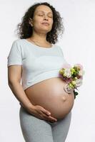 Delightful pregnant woman with bouquet of flowers, caressing her big belly in late pregnancy, isolated white background photo