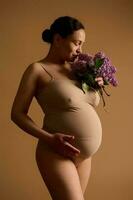hermosa embarazada mujer en beige lencería, participación mano en su grande barriga, oliendo un manojo de floreciente púrpura lilas foto