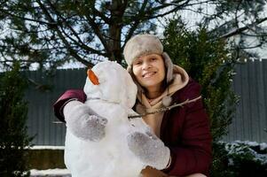 hermosa mujer en calentar invierno ropa y oído solapas sombrero, sonriente a cámara, abrazando un monigote de nieve en un nieve cubierto parque foto