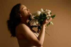 Beautiful multi-ethnic curly haired middle aged woman sniffing bunch of white blooming lilacs, isolated beige backdrop photo