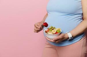 de cerca embarazada mujer en azul camiseta, participación un cuenco con delicioso Fruta ensalada terminado su grande barriga, aislado en rosado foto
