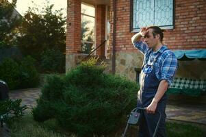 Male gardener in blue working uniform wiping sweat from forehead, tired after hard work in garden on hot summer day photo