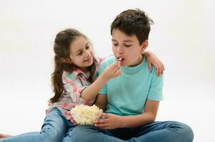 encantador pequeño niña abrazos y alimenta su hermano con sabroso palomitas de maiz mientras acecho película o dibujos animados terminado blanco antecedentes foto