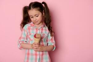 caucásico pequeño niño niña alegría a un delicioso hielo crema en gofre cono, aislado en rosado antecedentes foto