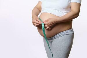 Gravid pregnant woman measuring her big tummy in the third trimester with a tape, isolated over white studio background photo