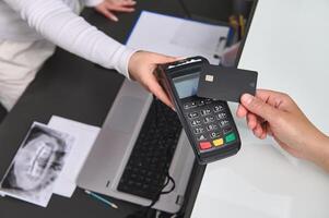 Top view customer hand holds black mockup credit card above a POS terminal, makes cashless payment using NFC technology photo