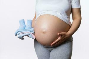Close-up pregnant gravid woman strokes her big belly in third pregnancy trimester, holds baby clothes isolated on white photo