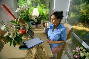 latín americano embarazada mujer tomando sorbo de café, sentado a mesa y trabajando en portapapeles en un floral diseño estudio foto