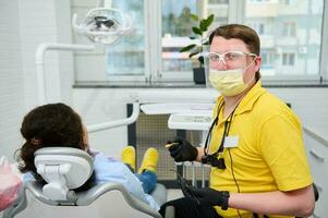 confidente dentista médico sonriente mirando a cámara, ejecutando dental tratamiento a un embarazada mujer en odontología clínica foto