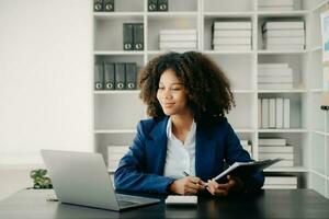 Beautiful African woman typing on tablet and laptop while sitting at the working wooden table modern office photo