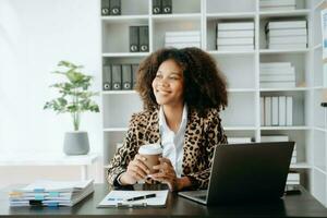 Young beautiful African woman typing on tablet and laptop while sitting at the working wooden table modern office photo