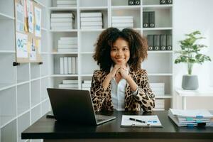 Young beautiful African woman typing on tablet and laptop while sitting at the working wooden table modern office photo