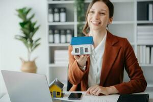 Young real estate agent worker working with laptop and tablet at table in office and small house beside it. photo