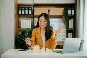 Asian Young real estate agent worker working with laptop and tablet at table in office and small house beside it. photo