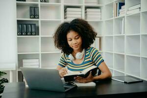 young African student studying at the college library, sitting at the desk, using a laptop computer, tablet and headphones having a video chat. photo