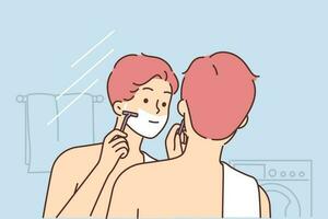 Man shaves face standing in bathroom and looking in mirror doing daily morning hygiene routine. Guy with naked torso and towel on shoulder stands with back to camera and shaves face going to work vector