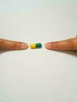 Isolated white photo of two forefingers pointing at a green and yellow medicine capsule.